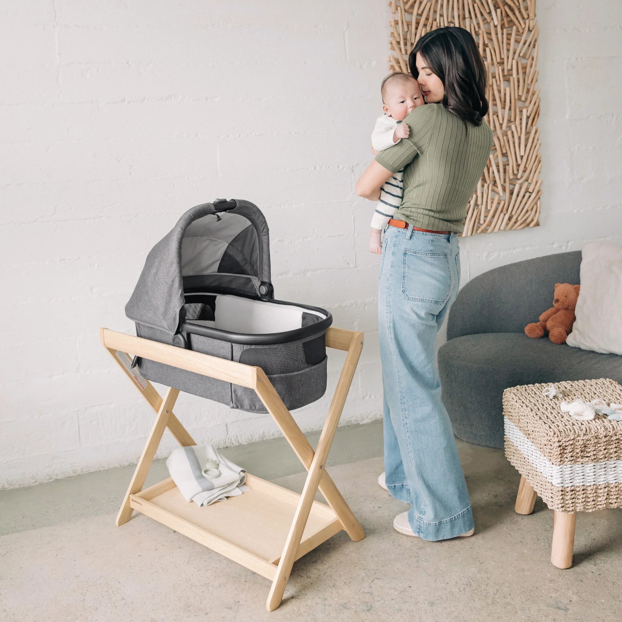 Mother holding newborn next to a grey bassinet on a wooden stand in a bright, modern nursery living space.