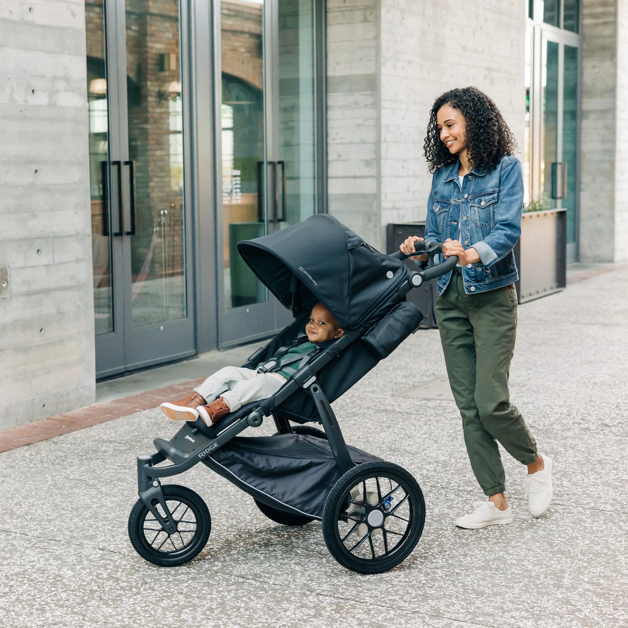 Woman pushing a black jogging stroller with a child inside, standing in front of a building.