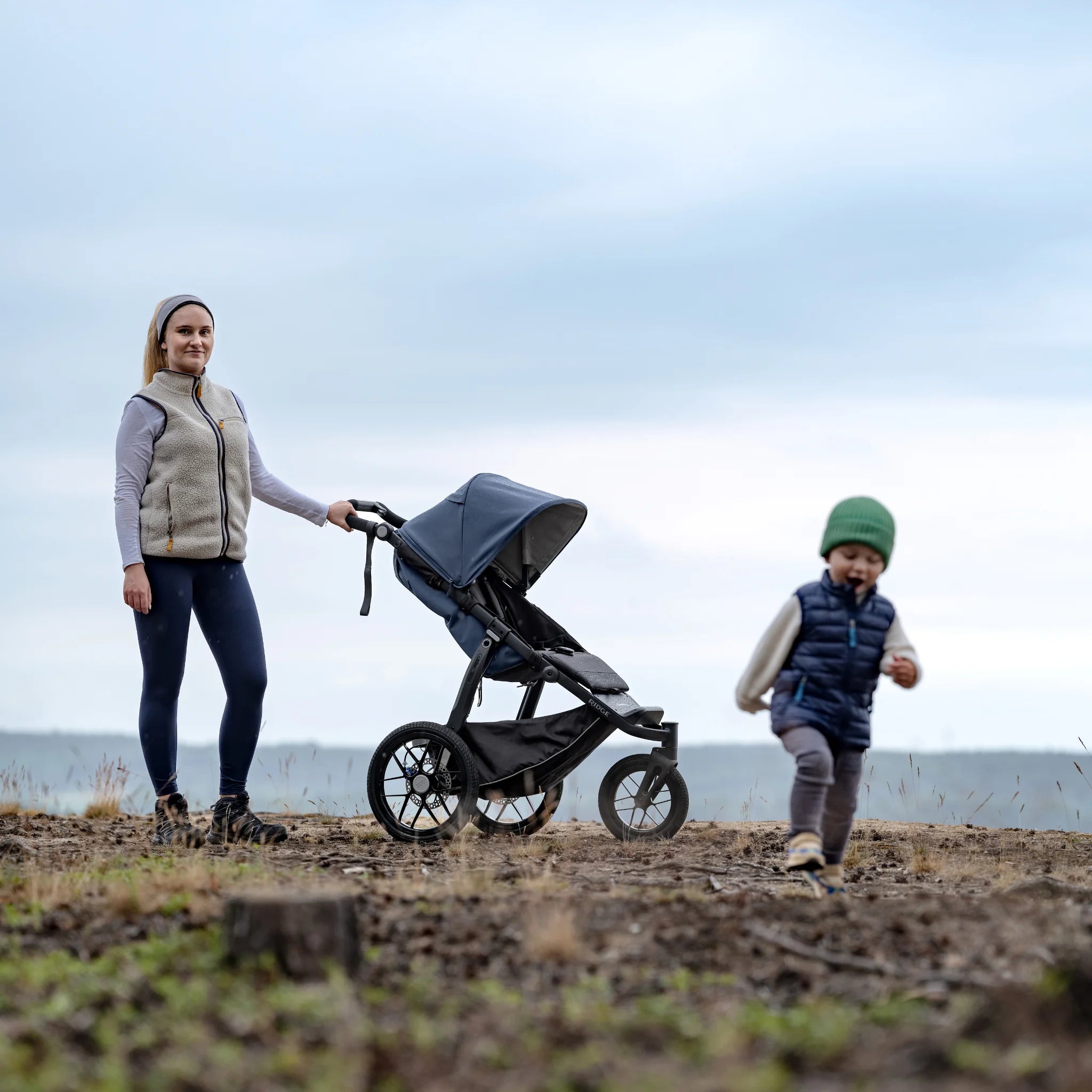 Woman pushing a three-wheel stroller with a child walking beside it in an open field.