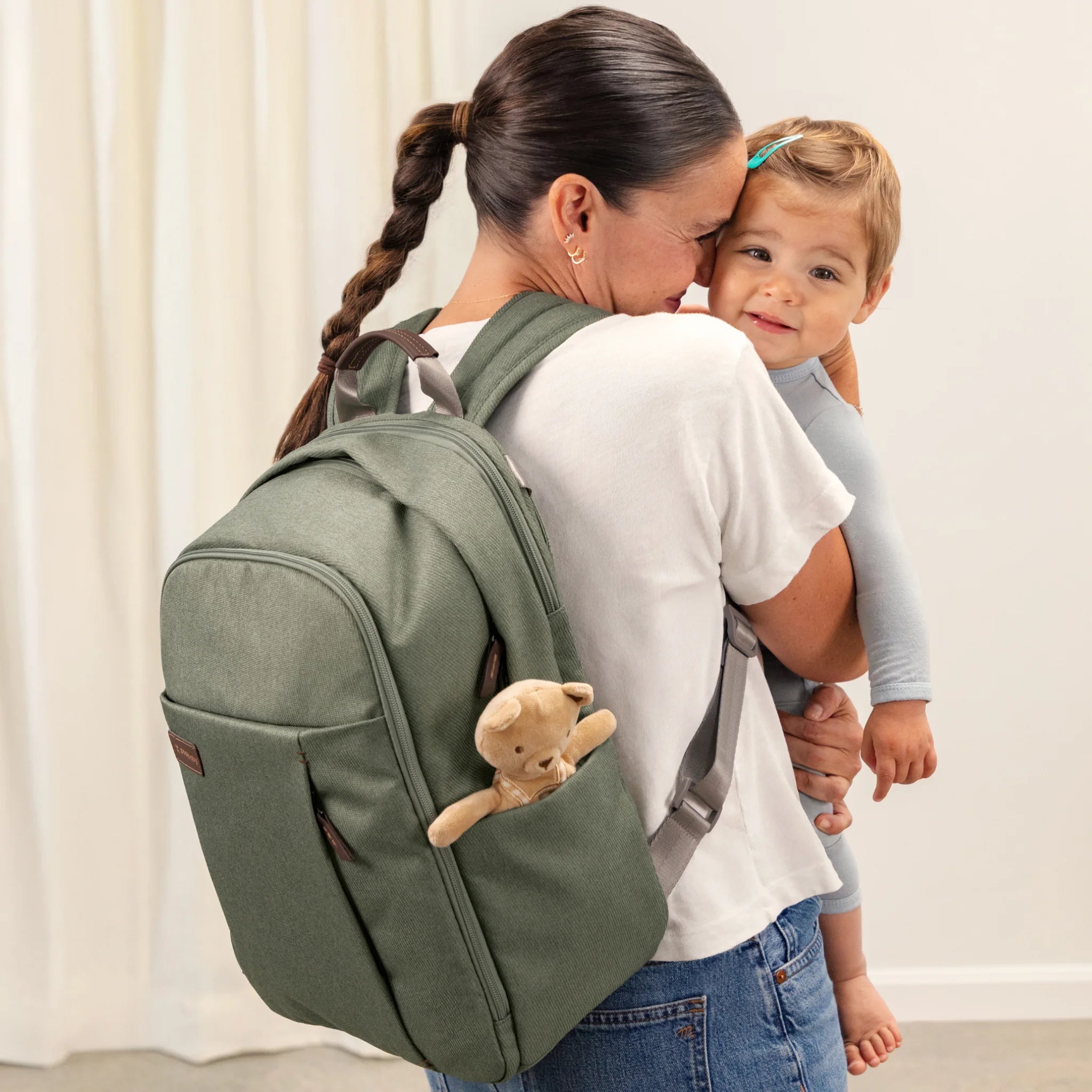 Woman holding a toddler while wearing a green UPPAbaby changing backpack; a small teddy bear peeks out of the backpackβs side pocket, with a light/white curtain background.