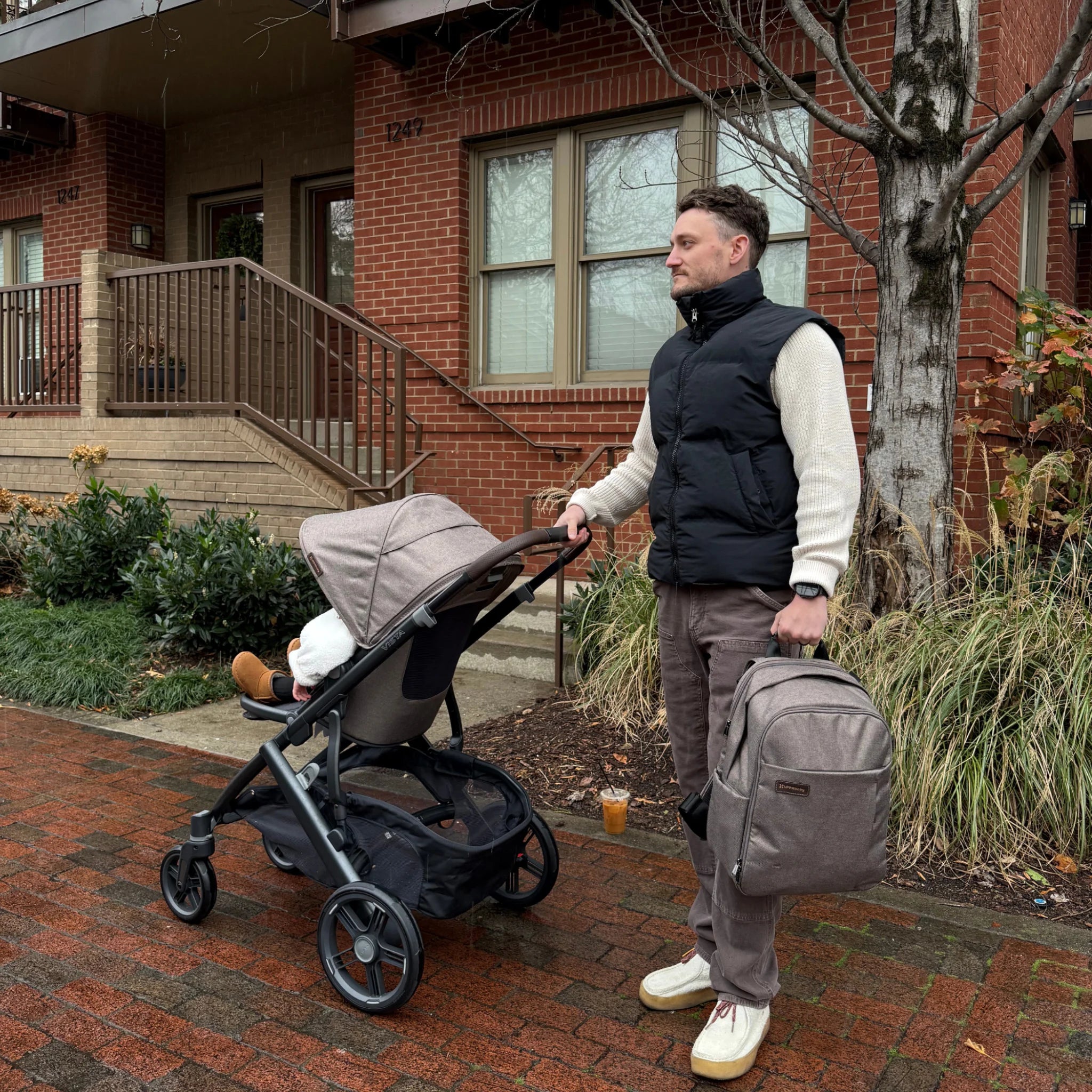 Adult pushing a stroller with a child inside while holding a brown UPPAbaby changing backpack on a wet sidewalk in front of a brick building.