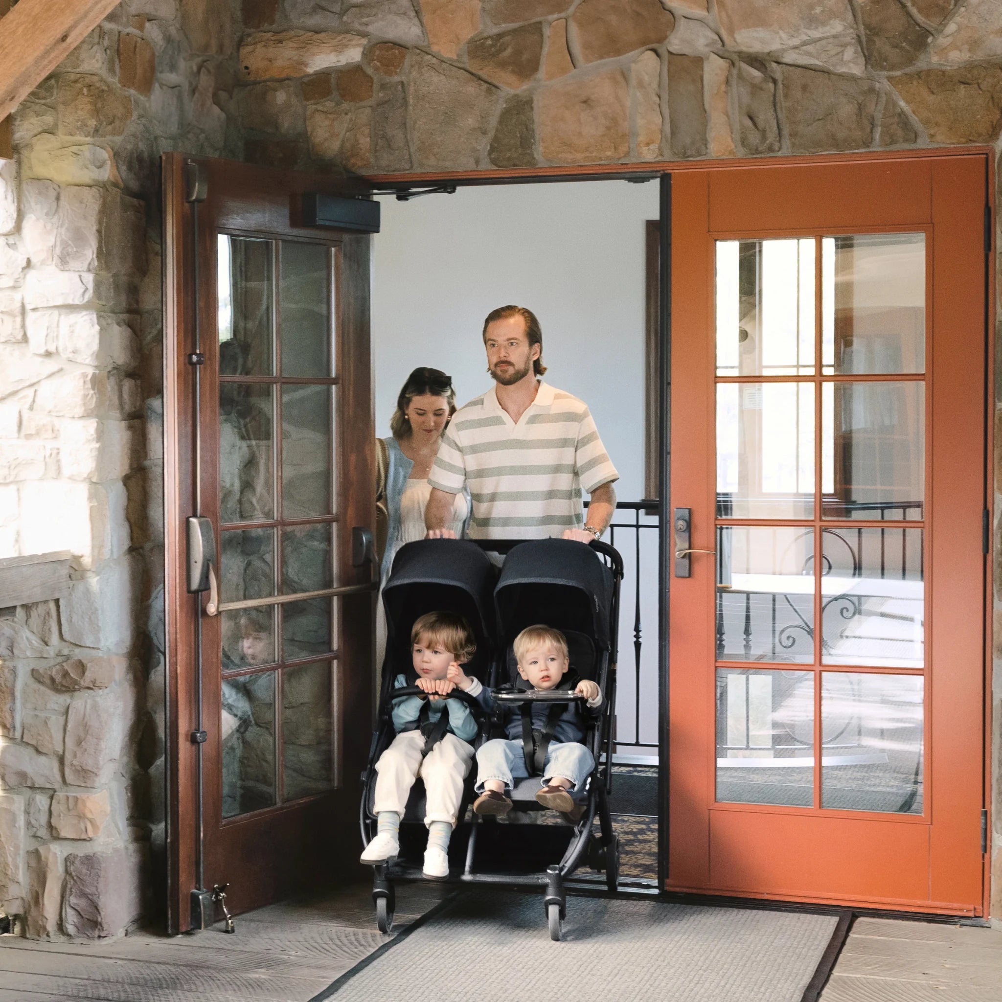 Parents pushing two children in a UPPAbaby Minu Duo double stroller with snack tray, exiting a stone building.