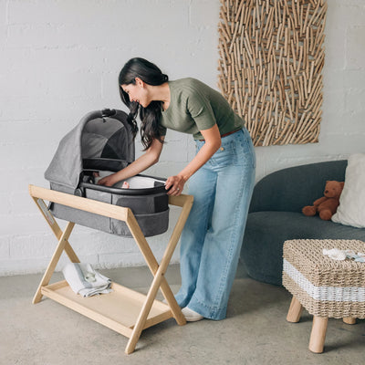 Woman setting up a grey UPPAbaby carrycot bassinet on a wooden stand in a modern living room.