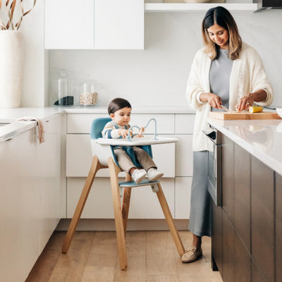 Child sitting in a blue Ciro high chair in a bright kitchen while an adult prepares food at the counter.