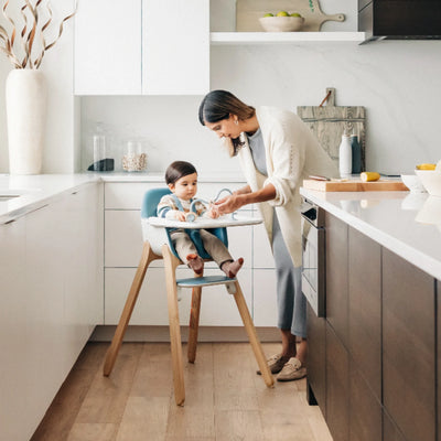 Adult helping a child at the tray of a blue Ciro high chair in a bright, modern kitchen.