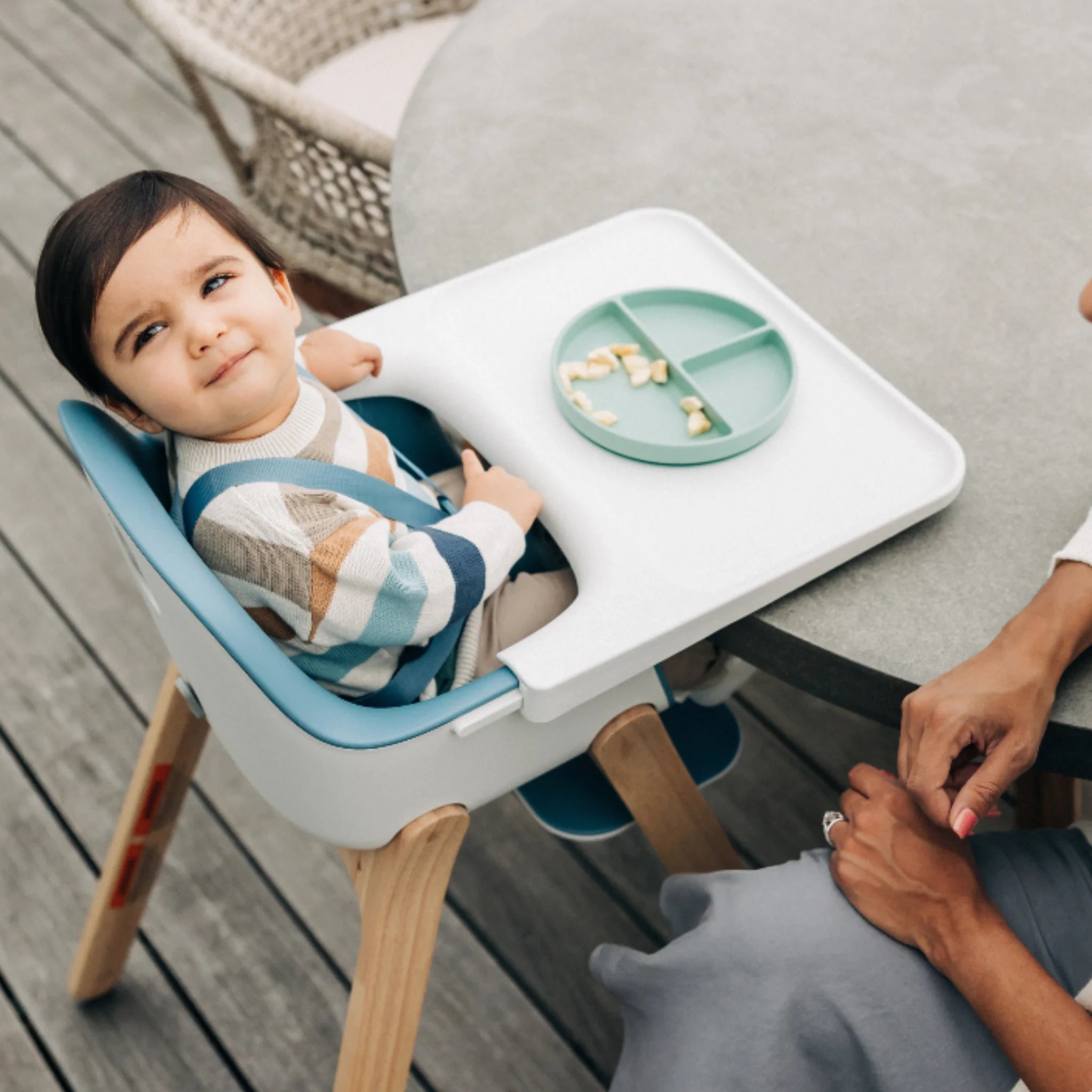 Child sitting in a blue Ciro high chair with a white tray and divided plate on an outdoor patio.