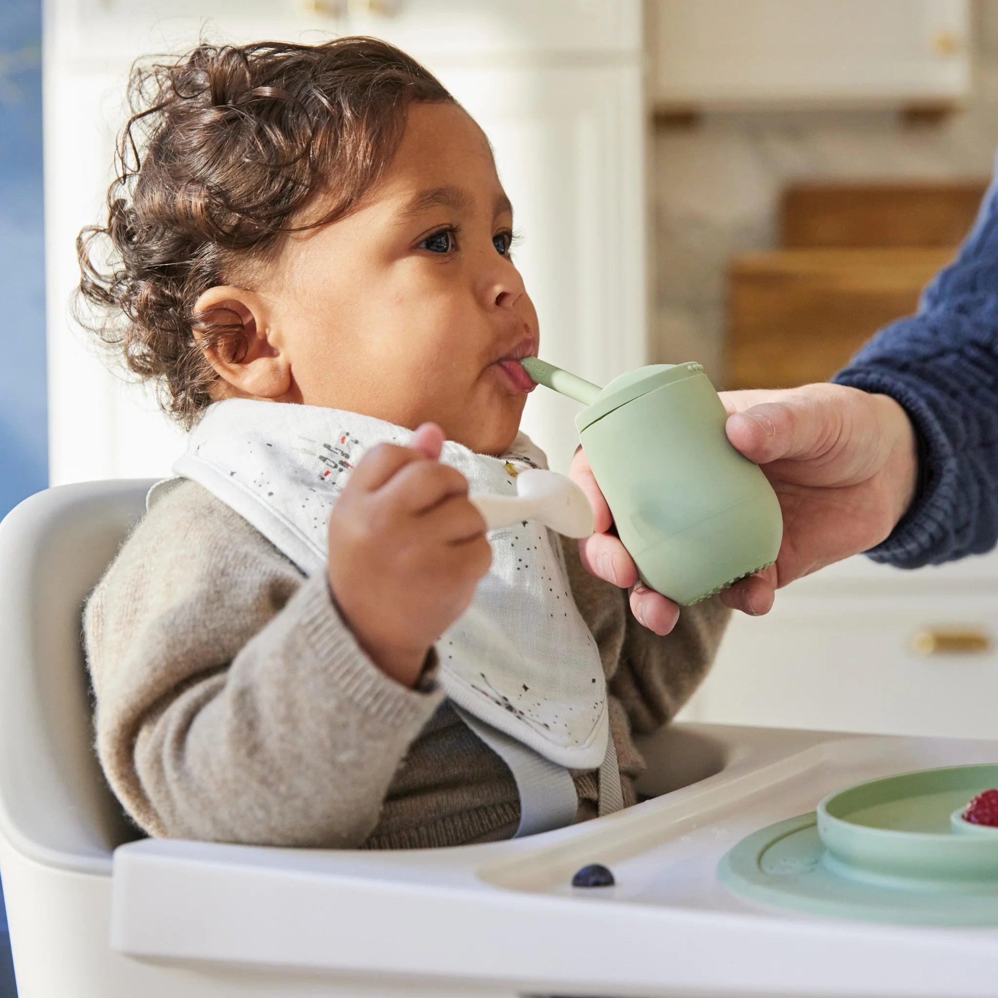 Toddler sitting in a Ciro high chair while an adult holds a green sippy cup to their mouth in a kitchen.