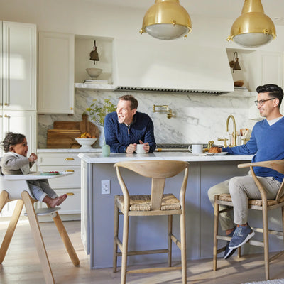 Toddler sitting in a Ciro high chair while two adults talk and smile at a kitchen island in a bright, modern kitchen.
