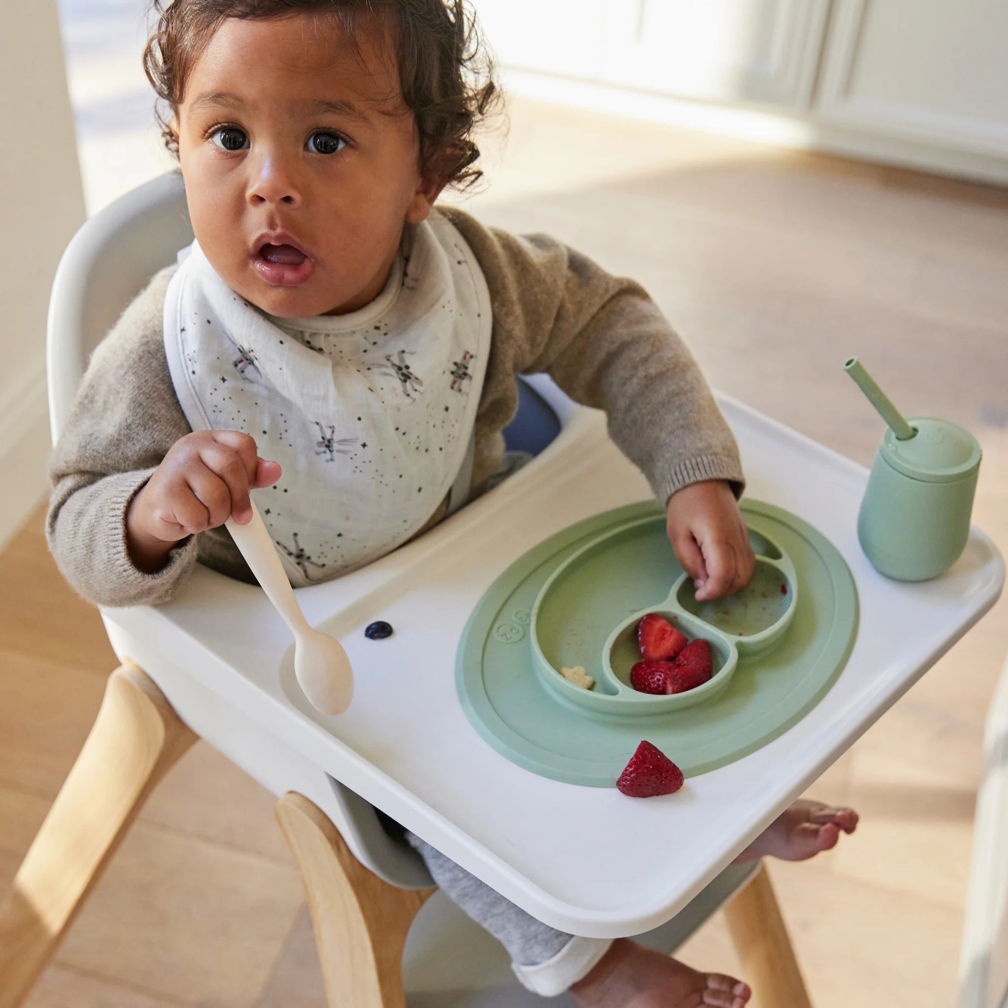 Toddler seated in the Ciro high chair with tray, holding a spoon beside a plate and cup on a wooden floor
