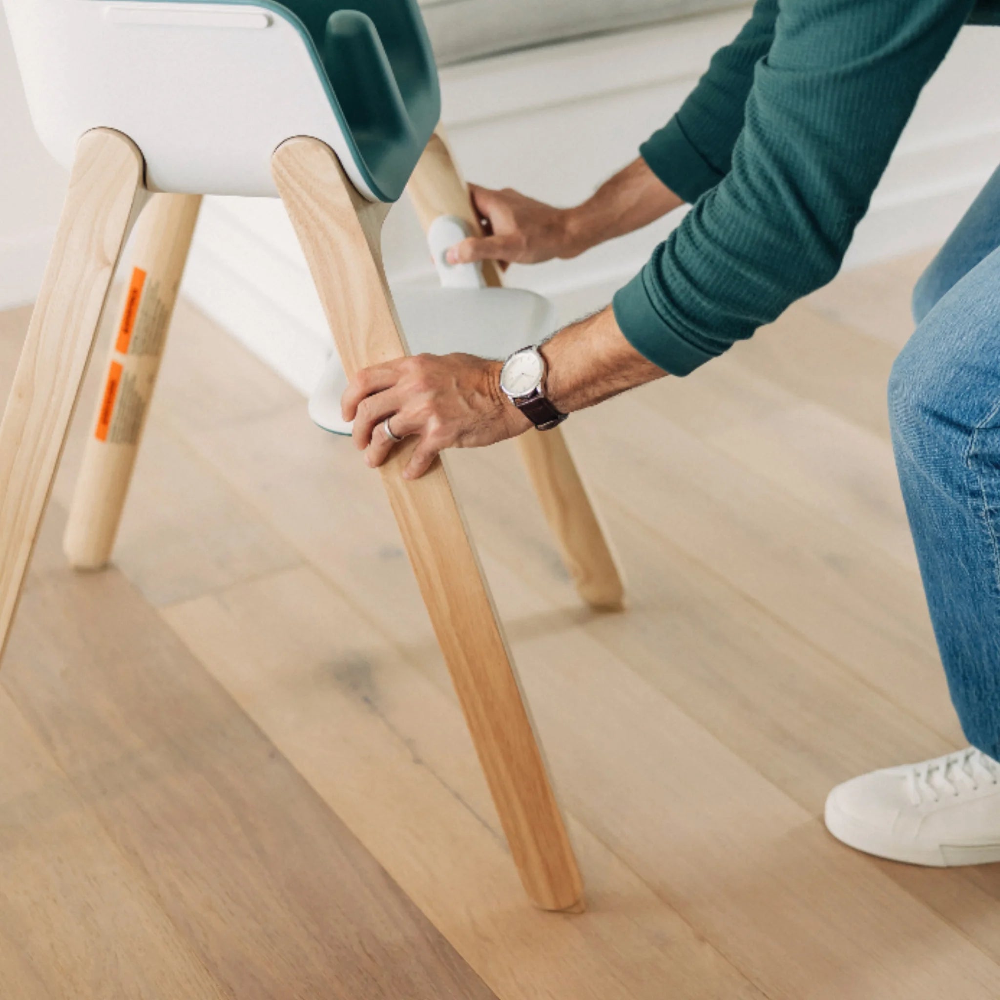 Adult adjusting the footrest on the Emrick Ciro high chair with wooden legs on a light wood floor.