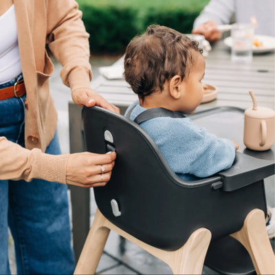 Adult adjusts the back strap on the Jake Ciro high chair while a child sits at an outdoor table.