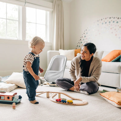 Parent and toddler playing at home with the UPPAbaby Mira bouncer in Stella in the background