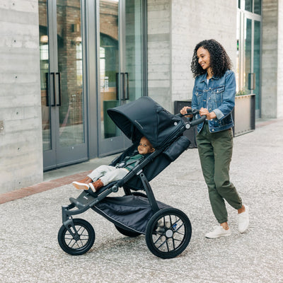 Woman pushing a black jogging stroller with a child inside, standing in front of a building.