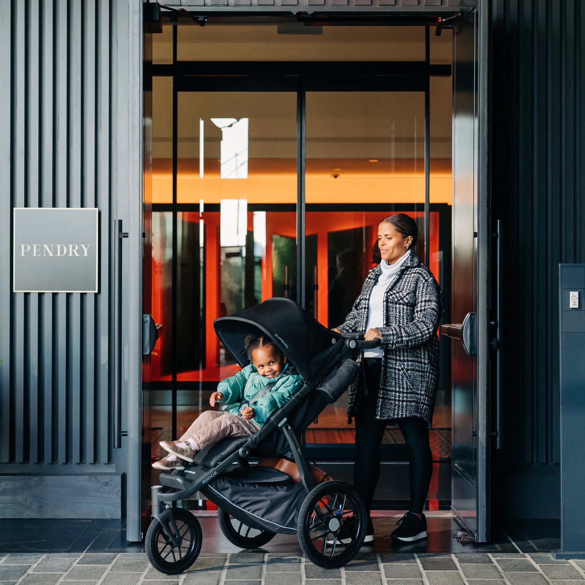 Woman pushing a stroller with a child in front of a building entrance.