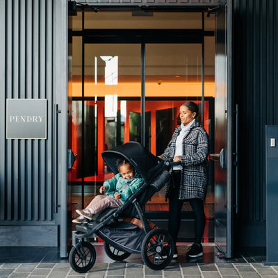 Woman pushing a stroller with a child in front of a building entrance.
