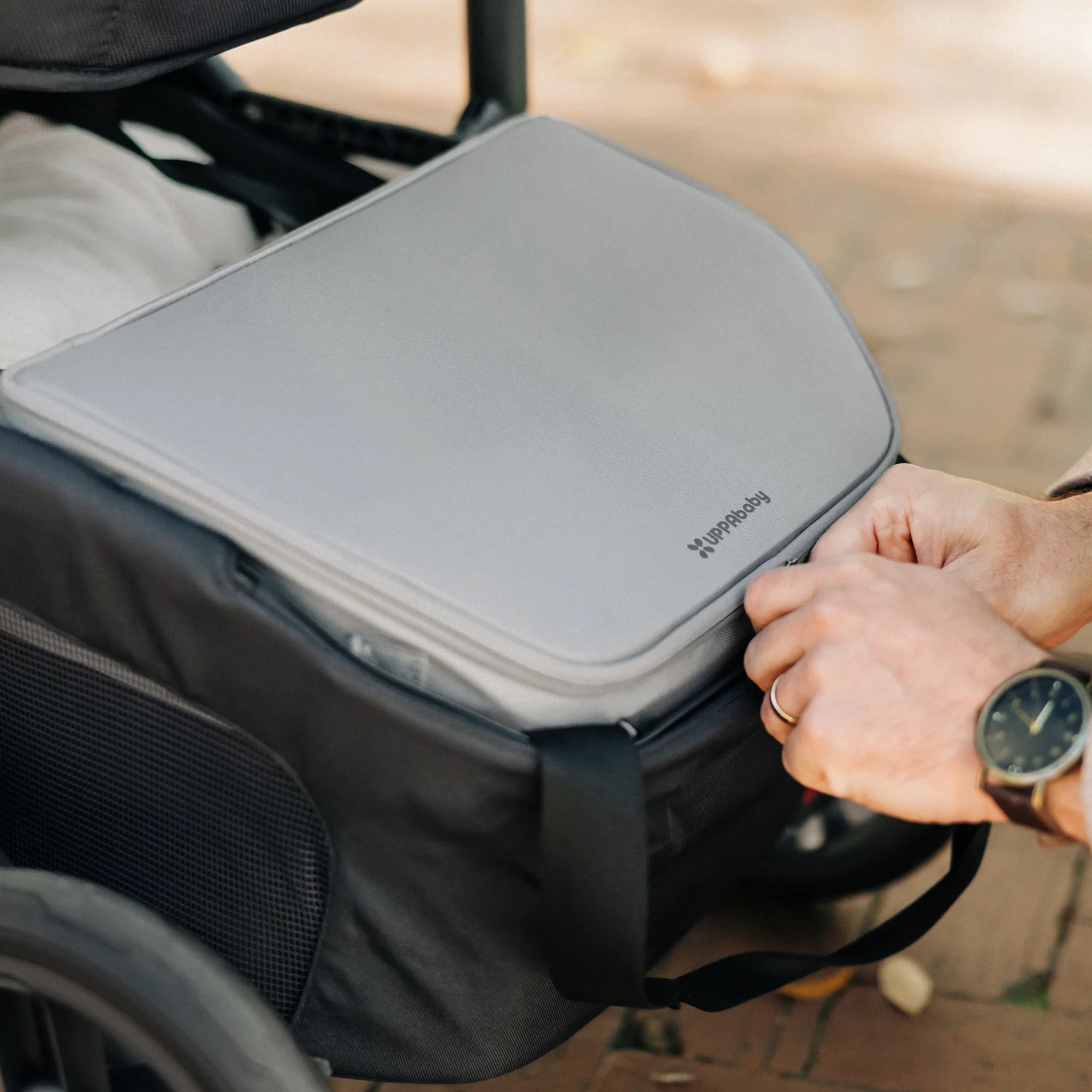 Close-up of hands placing a grey UPPAbaby Bevvy cooler bag into the under-seat basket of a stroller outdoors.