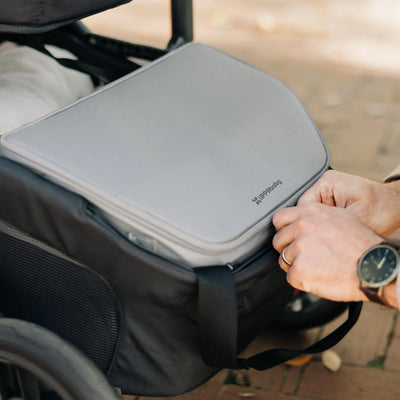 Close-up of hands placing a grey UPPAbaby Bevvy cooler bag into the under-seat basket of a stroller outdoors.