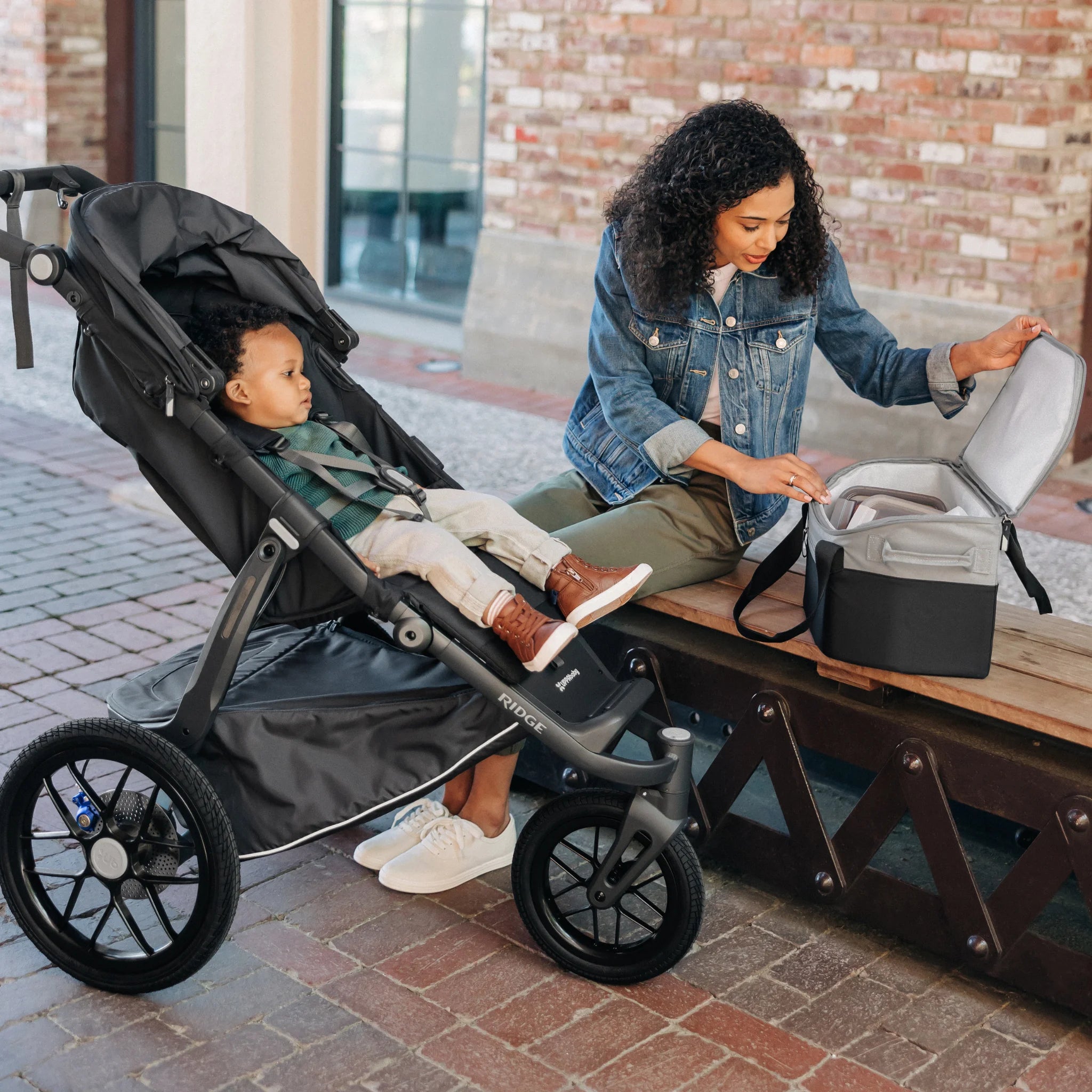 Woman sitting beside a stroller with a child inside, opening a grey and black UPPAbaby Bevvy cooler bag on a bench outdoors.