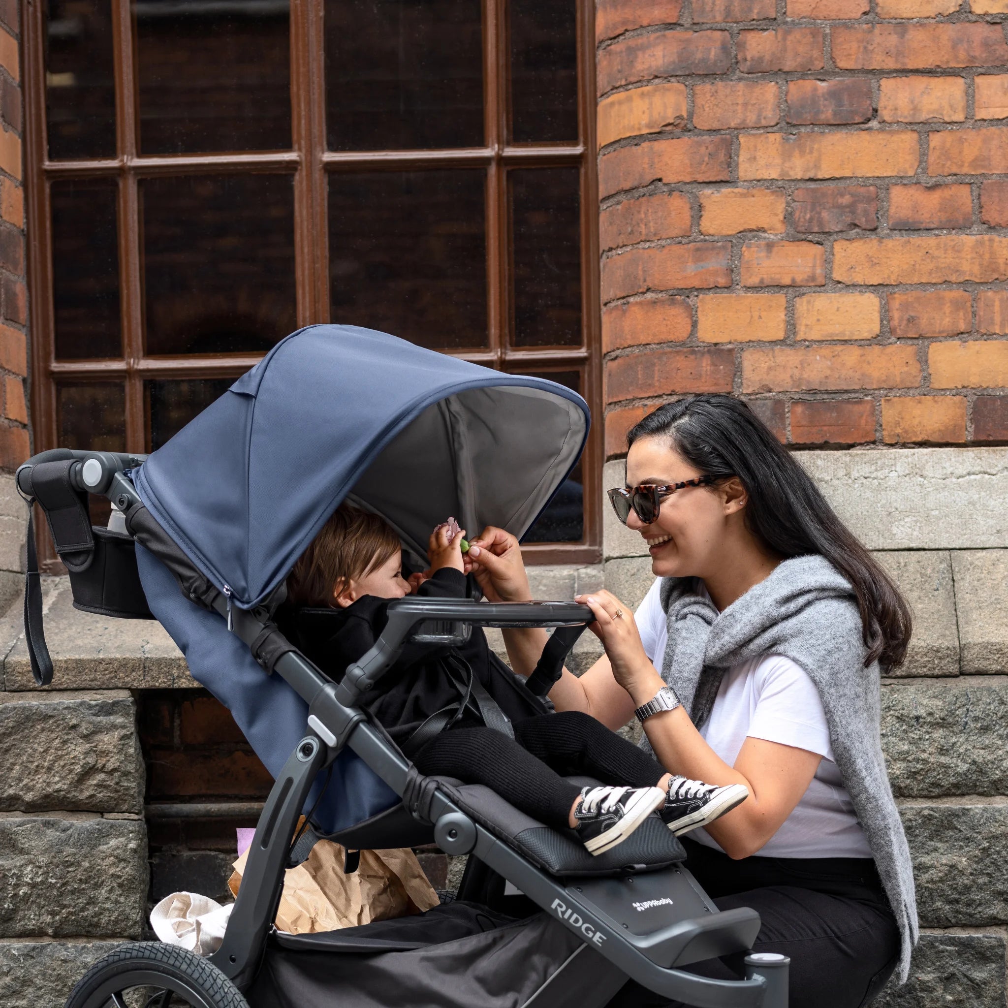Woman sitting beside a stroller with a navy canopy, smiling and feeding a child seated inside, with the snack tray attached to the stroller, set against a brick wall background.