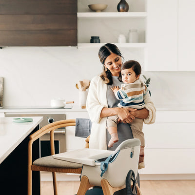 Mom holding and smiling at baby in the kitchen, infront of high chair with high chair cushion accessory. 
