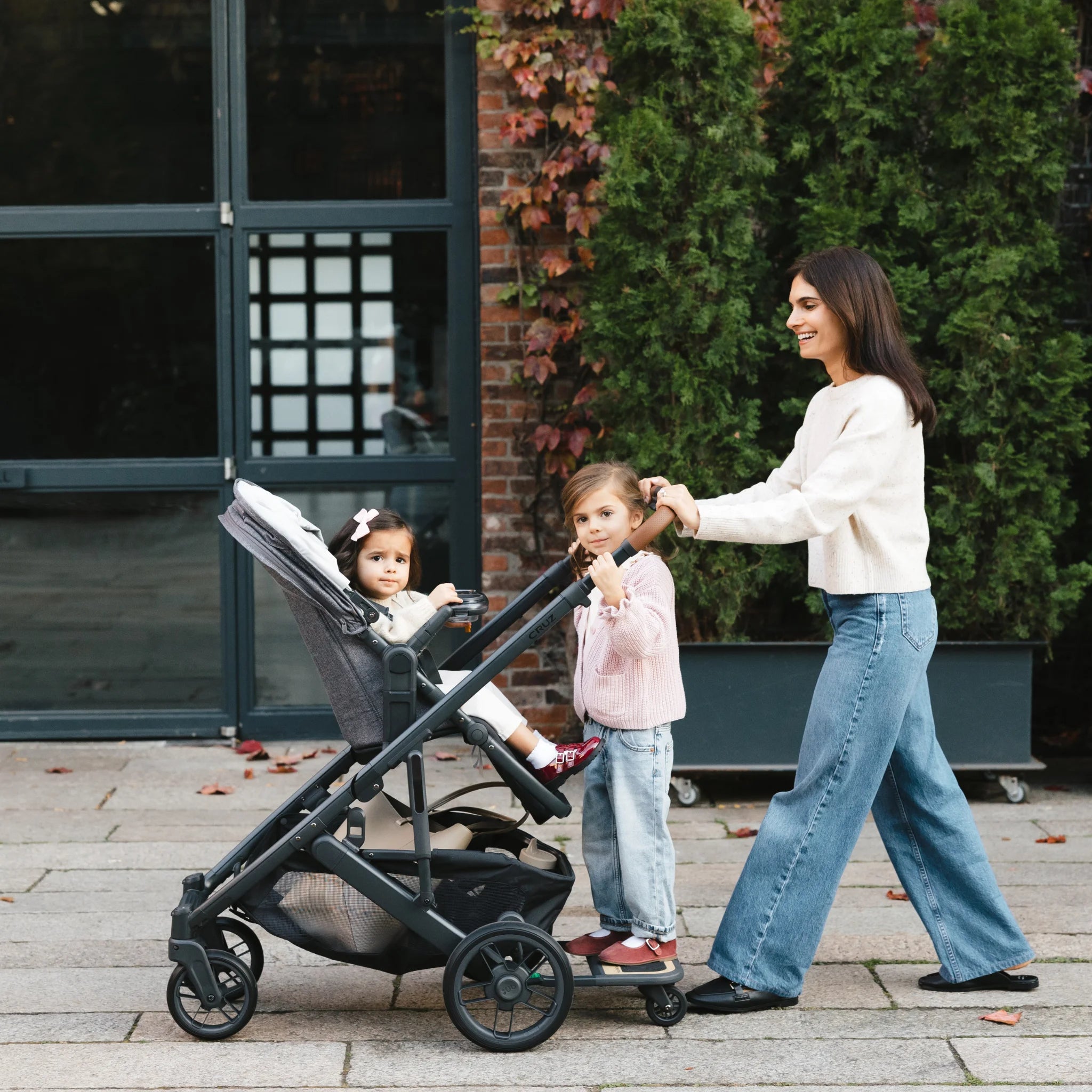Mom laughing and pushing cruz stroller with one kid sitting and one kid standing on cruz piggyback board. 