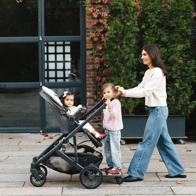 Mom laughing and pushing cruz stroller with one kid sitting and one kid standing on cruz piggyback board. 