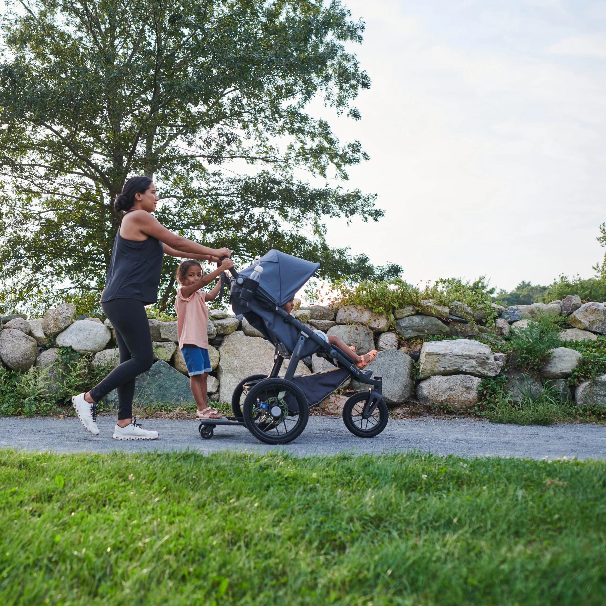 Mum walking and pushing ridge stroller outside, with one kid standing on the ridge piggyback board. 