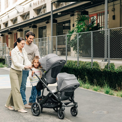 Parents smiling at vista stroller, pushing three kids outdoors. 