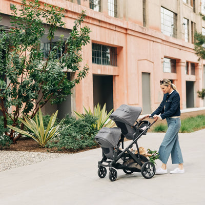 Mum pushing and smiling at double grey rumbleseat stroller outdoors. 